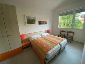 Ein Schlafzimmer mit einem Bett, zwei Stühlen, einem Kleiderschrank, einem Nachttisch, zwei Lampen und einem Fenster mit Blick auf Grünflächen.