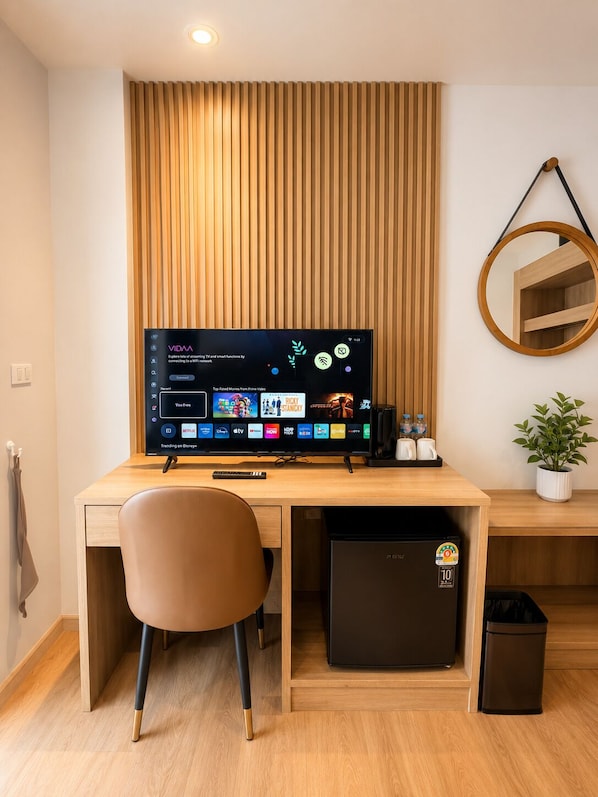 A modern hotel room with a wooden desk, a computer, a chair, a small fridge, a trash bin, a potted plant, and a wall-mounted mirror. - Azizi Boutique Hotel (Patong)