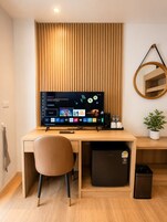 A modern hotel room with a wooden desk, a computer, a chair, a small fridge, a trash bin, a potted plant, and a wall-mounted mirror.