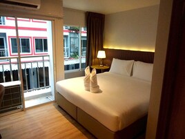 Habitación de hotel con cama, mesita de noche, lámpara, ventana con cortinas y vista a edificios.