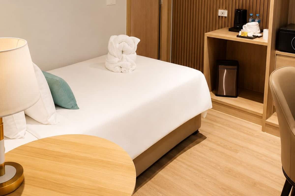 A hotel room with a bed, a bedside table with a lamp, a chair, a wooden wardrobe, and a small shelf with toiletries.
