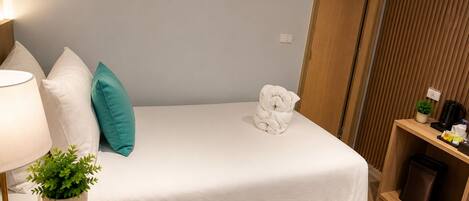 A hotel room with a bed, a bedside table with a lamp and a plant, a wooden shelf with a coffee maker, and a door.