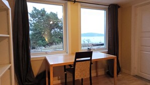 A room with a wooden table and chair, a window with curtains, and a view of trees and water. - Sea Cottages 10 - The First Seaview Apartment (Åkersberga)