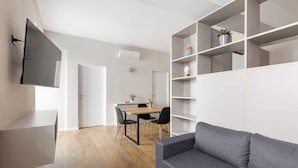 A modern living room with a grey sofa, a TV mounted on the wall, a dining area with a table and chairs, and a white shelving unit. - 2 min to Lake Maggiore | Modern & Central (Cannobio)