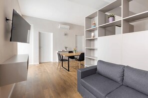 A modern living room with a grey sofa, a TV mounted on the wall, a dining area with a table and chairs, and a white shelving unit. - 2 min to Lake Maggiore | Modern & Central (Cannobio)
