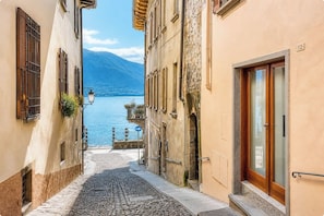 A narrow cobblestone street with buildings on either side, a mountain in the background, and a body of water. - 2 min to Lake Maggiore | Modern & Central (Cannobio)