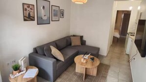 Un salon moderne avec un canapé d’angle gris, une table basse ronde en bois et un luminaire suspendu en fibres tressées. On y trouve des tableaux encadrés au mur, une petite table d’appoint en bois avec une plante, et un sol carrelé.