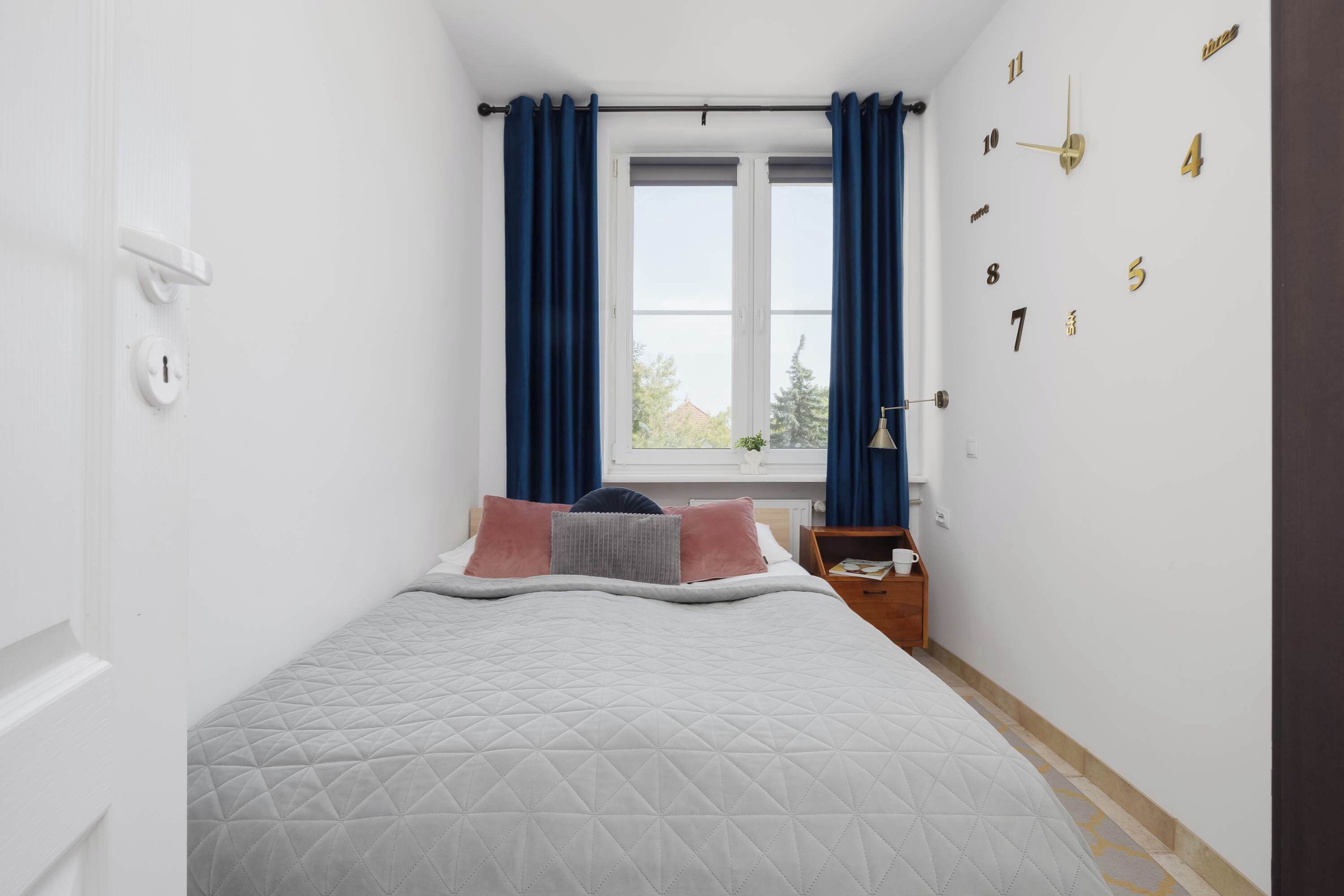 Un dormitorio con una cama, una ventana con cortinas, un reloj de pared y una mesita de noche con una lámpara.