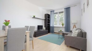 A modern living room with a dining table, grey chairs, a grey sofa, a black entertainment unit, and a window with blue curtains. - Cozy Apartment for Friends | Great Location Close to the Metro (Warszawa)