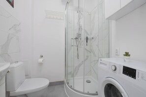 A modern bathroom with a toilet, a glass-enclosed shower, and a front-loading washing machine. - Cozy Apartment for Friends | Great Location Close to the Metro (Warszawa)