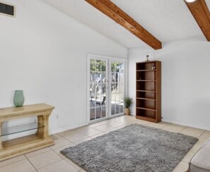 Ein Zimmer mit Holzbalkendecke, einer Glastür, die nach draußen führt, einem Holzregal und einem grauen Teppich.