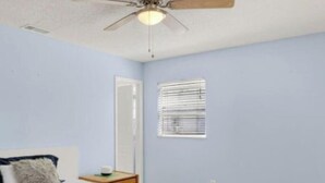 Ein Schlafzimmer mit einem Bett, einem Deckenventilator, einem Fenster mit Jalousien und einem Nachttisch.