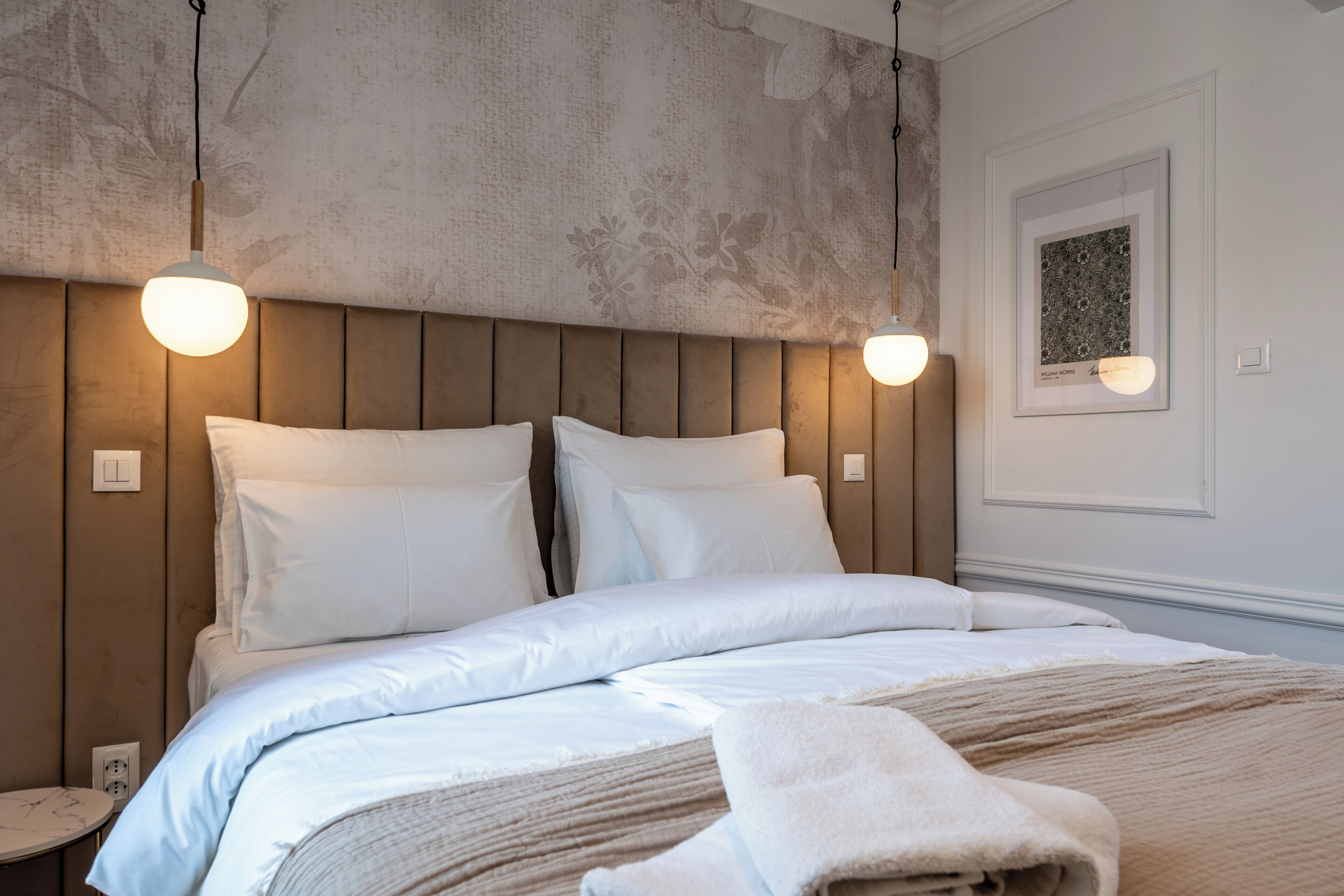 Een net opgemaakt bed met witte lakens, een houten hoofdbord en twee hanglampen.