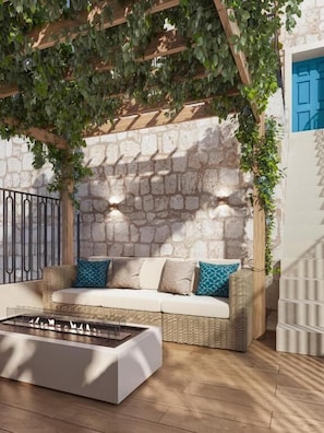 Un muro de piedra, un sofá de mimbre con cojines, un hogar y una pérgola de madera con plantas.
