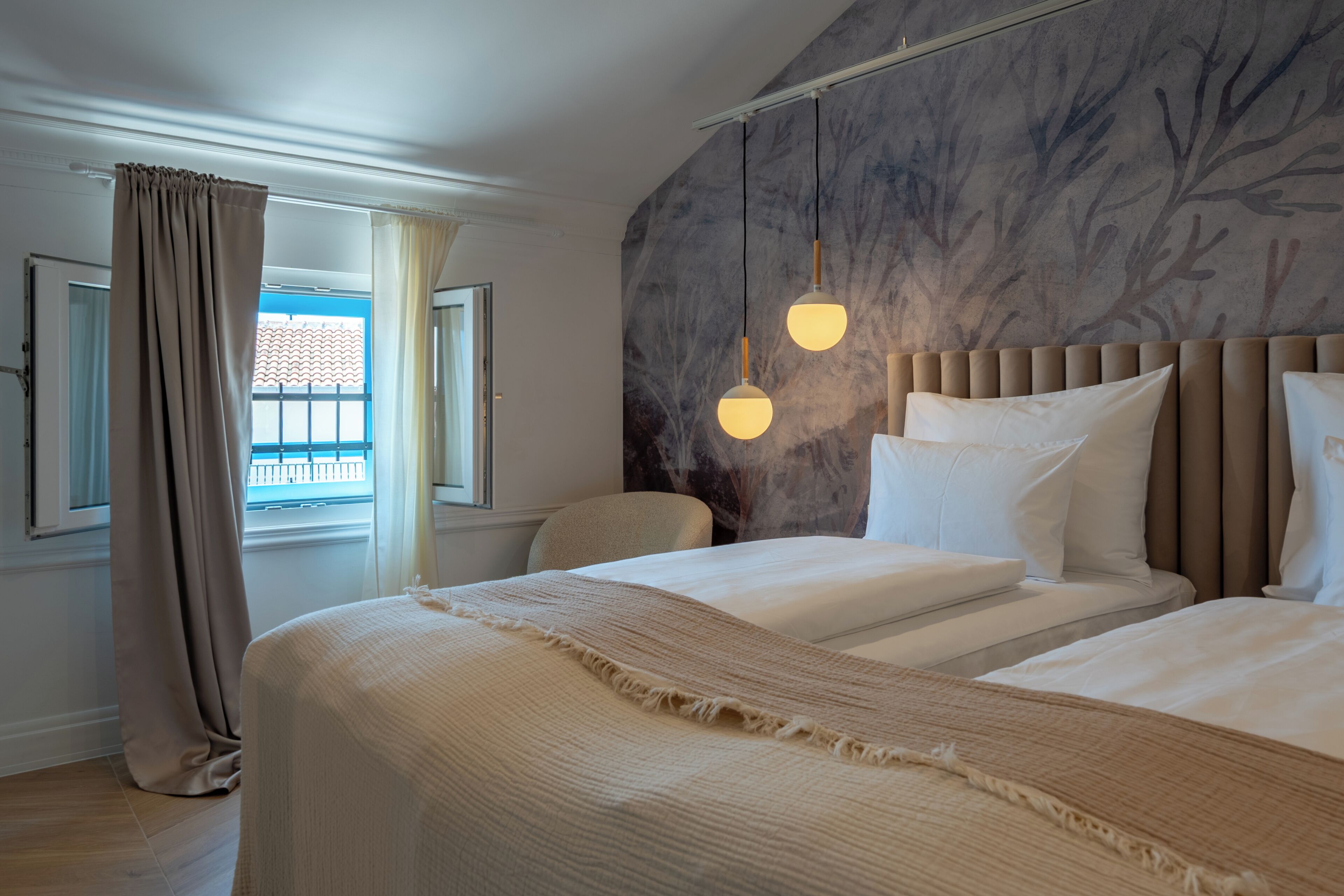 Habitación de hotel con cama, una silla, ventana con cortinas y un papel tapiz con estampado de árbol.