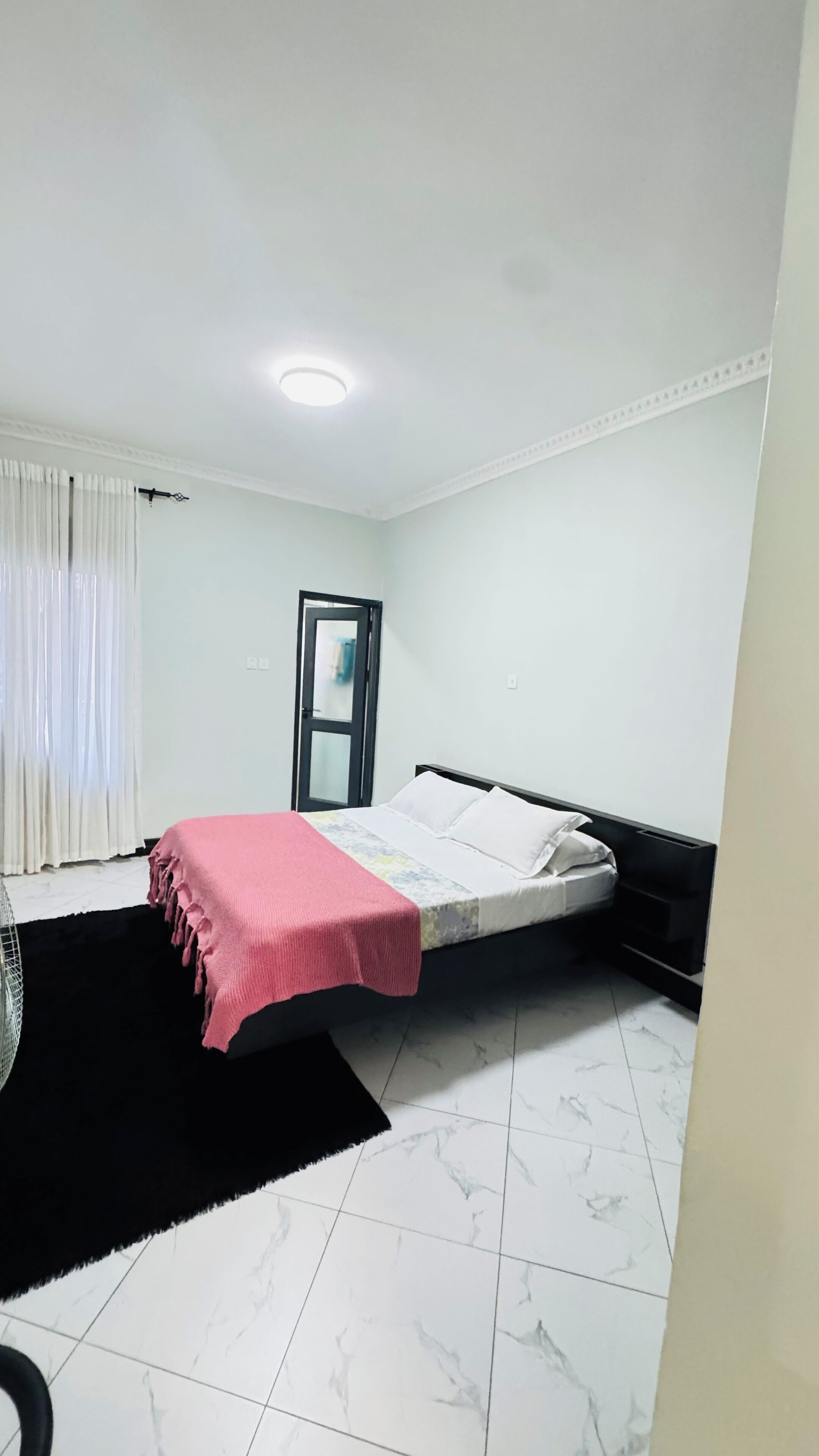 A bedroom with a bed, a black and white tiled floor, a white door, and a window with white curtains.