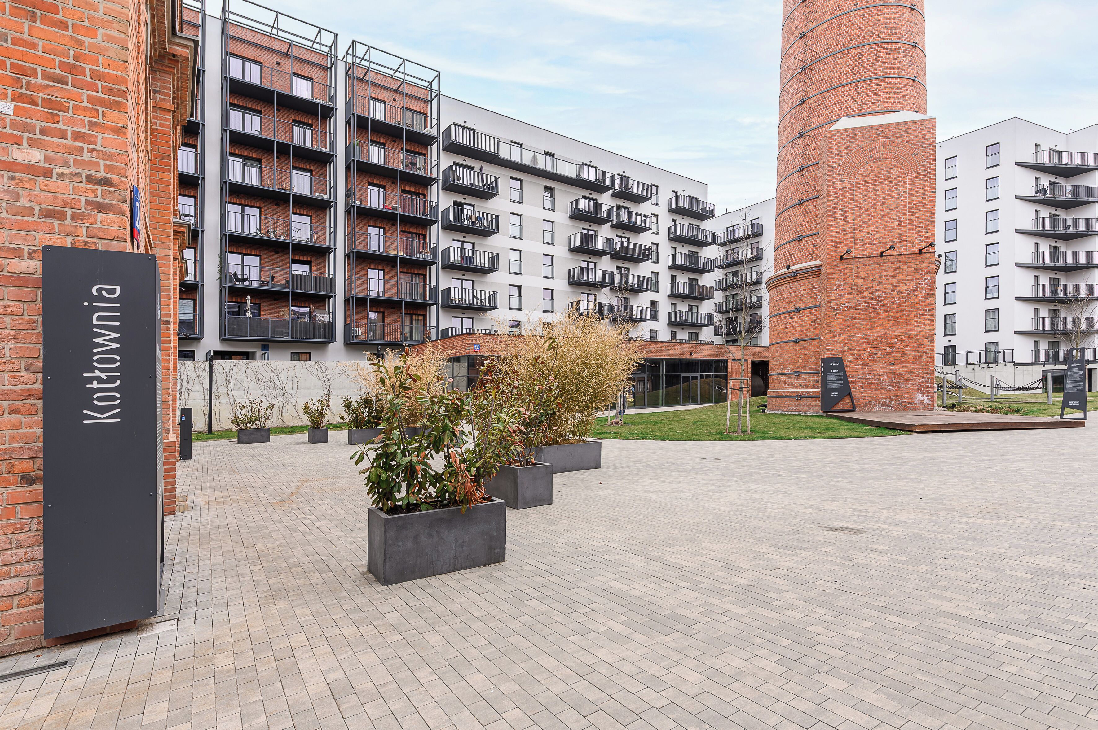 Un bâtiment moderne doté de nombreux balcons, une tour en briques et une enseigne indiquant « Kothownia ».