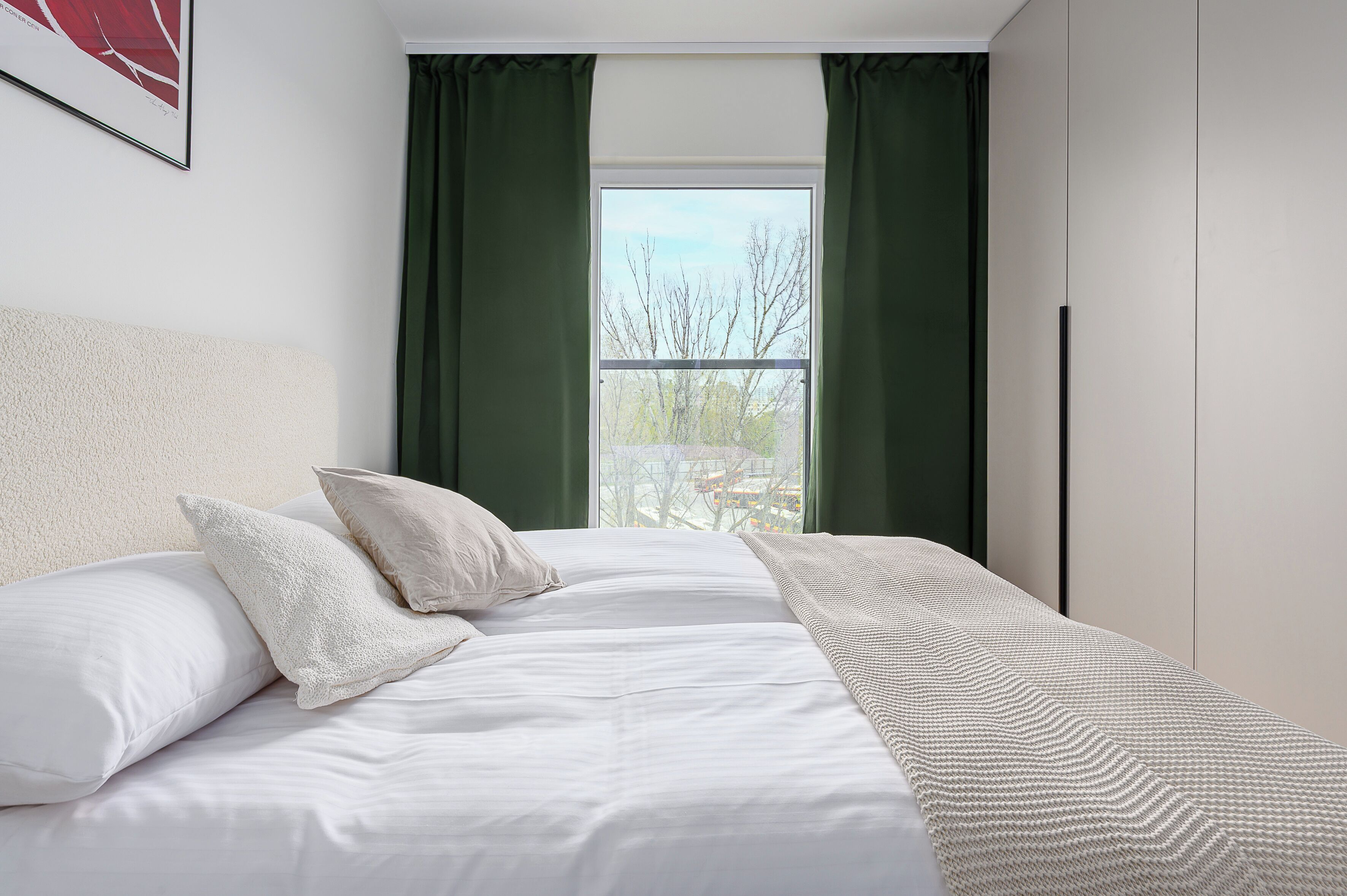 Quarto com cama, almofadas, janela com cortinas e vista para árvores.