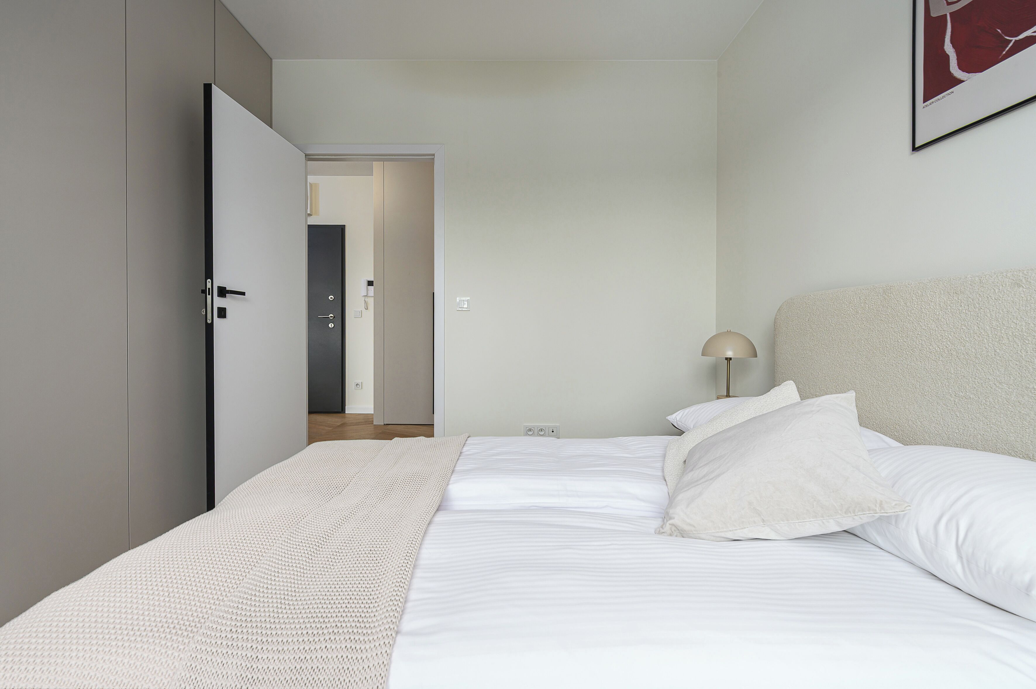 A bedroom with a bed, pillows, a bedside lamp, a wardrobe, and a door leading to another room.