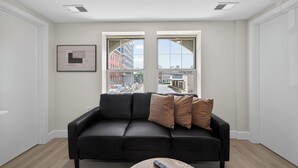 Un soggiorno moderno con un divano in pelle nera, due finestre con vista sulla città, un quadro appeso e un telecomando su un tavolino da caffè.