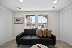 A modern living room with a black leather sofa, two windows with a city view, a framed picture on the wall, and a remote on a coffee table. - Bright and stylish fully furnished unit (Reading)