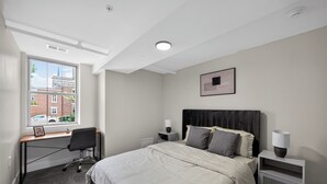 Un dormitorio con cama, escritorio, silla, mesitas de noche y ventana con vista a edificios.