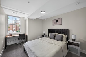 Ein Schlafzimmer mit Bett, Schreibtisch, Stuhl, Nachttischen und einem Fenster mit Blick auf Gebäude.