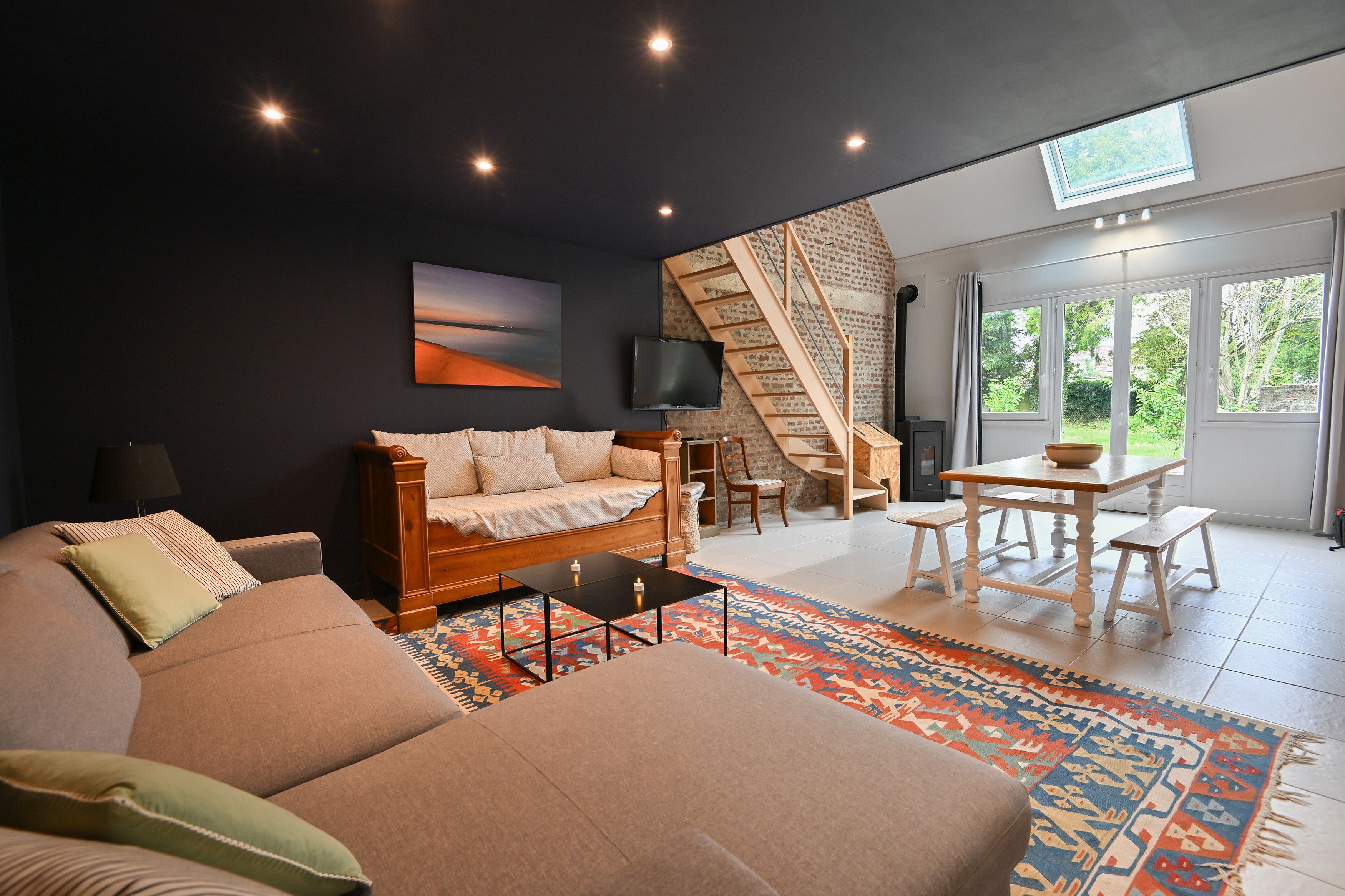 En moderne stue med sofa, sofabord og TV. Der er en trappe til en etage ovenover, og et stort vindue med udsigt over naturen.