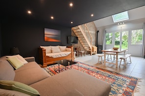 Un salon moderne avec un canapé, une table basse et un téléviseur. Un escalier mène à un niveau supérieur, et une grande fenêtre offre une vue sur l’extérieur.