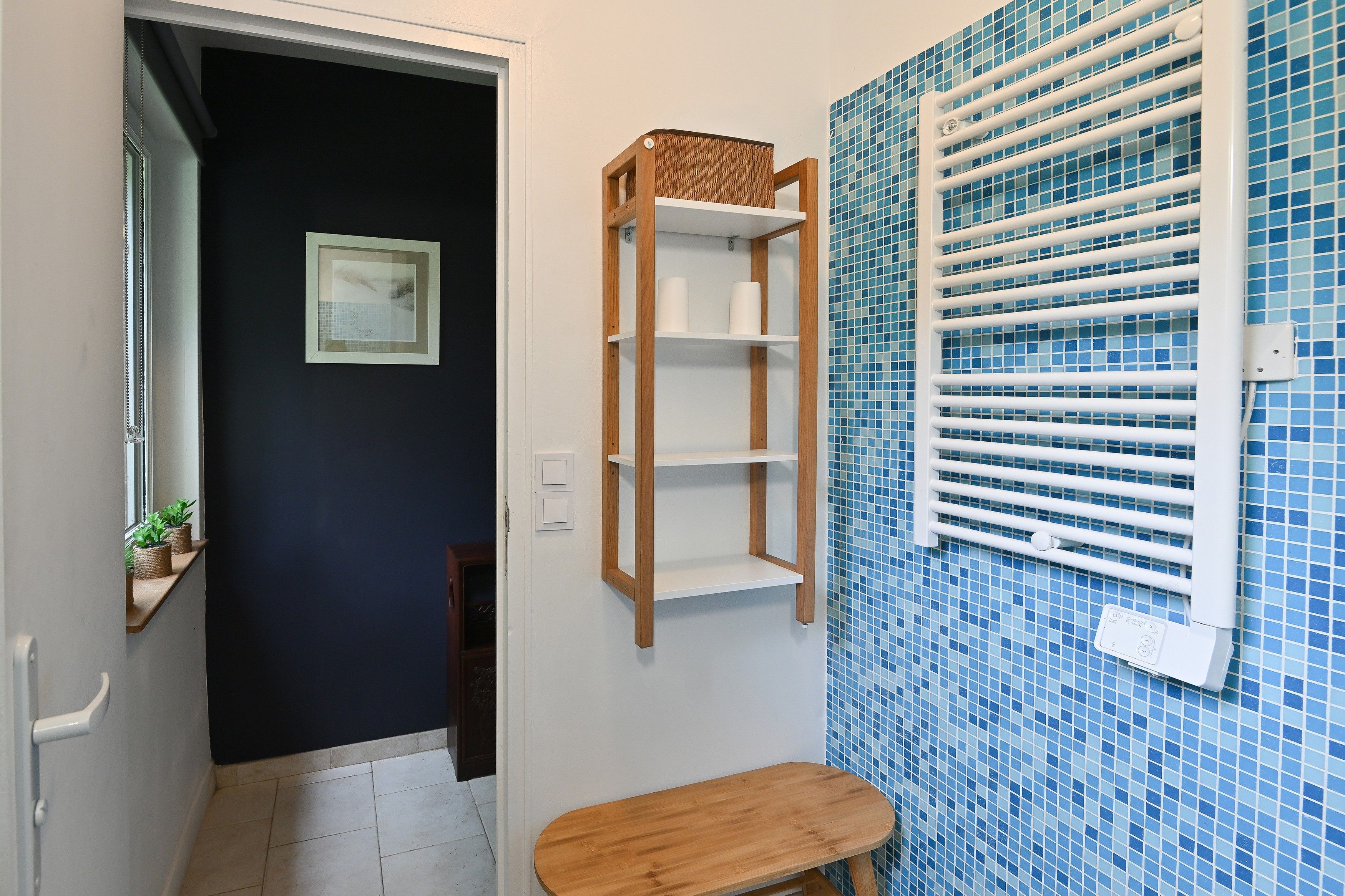 Ein Badezimmer mit einem hölzernen Regal, einer weißen Toilette, einer blauen Fliesenwand und einem Fenster mit Jalousien.