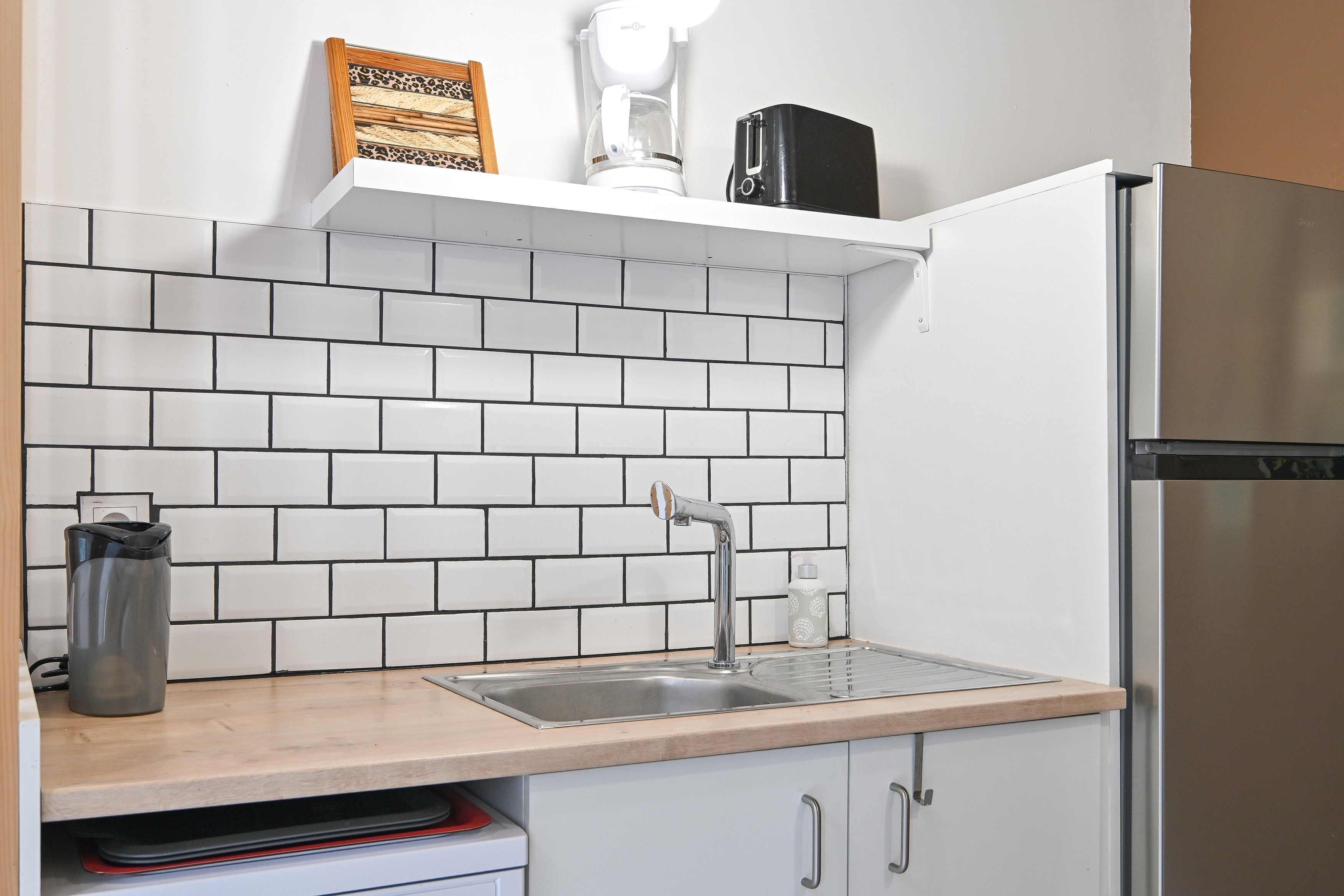 Une cuisine avec des carreaux de céramique blancs disposés en motif métro, un évier en acier inoxydable, une machine à café et un réfrigérateur.