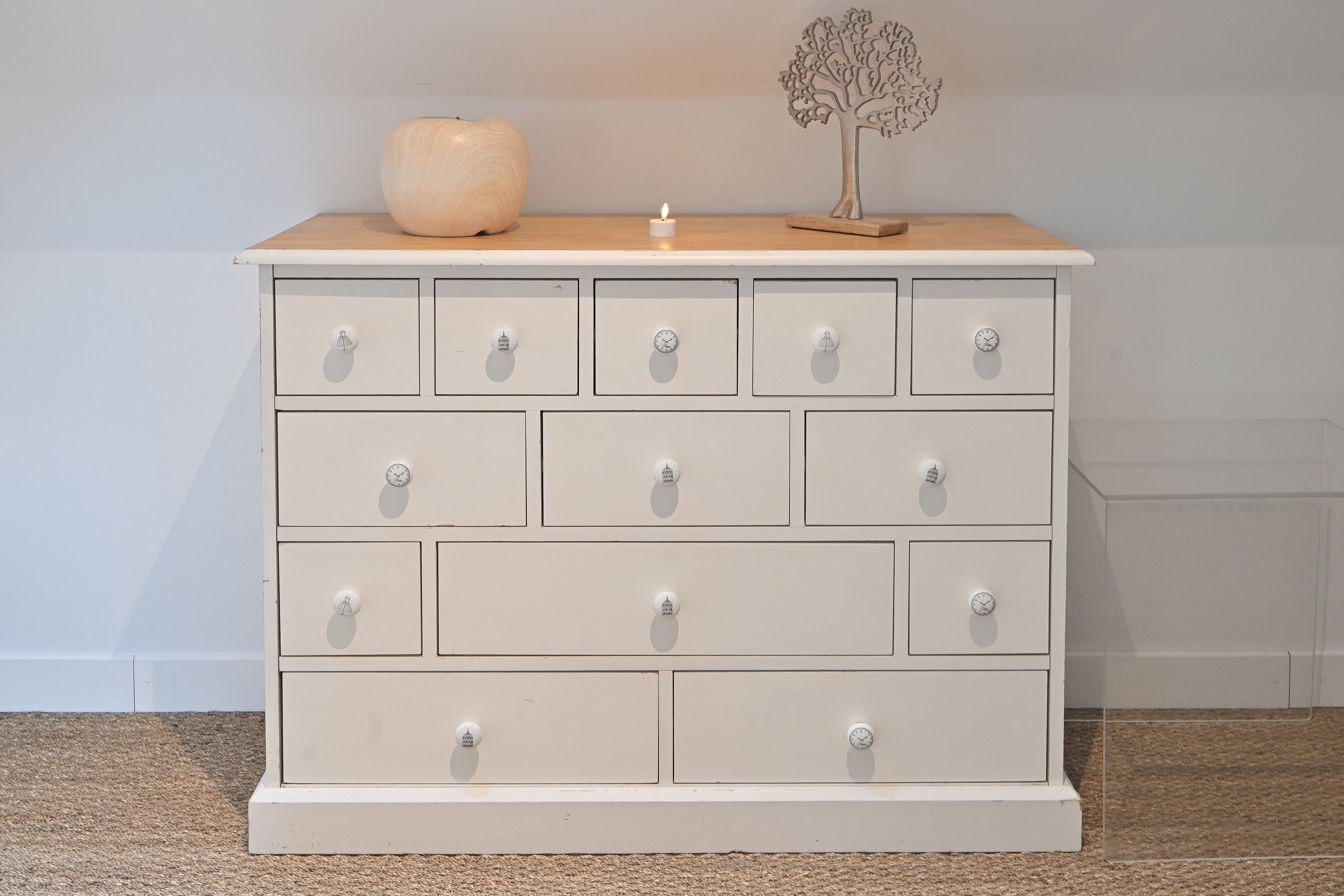 Een witte ladekast met meerdere laden en knoppen, een kleine houten decoratie erop, en een kaars.