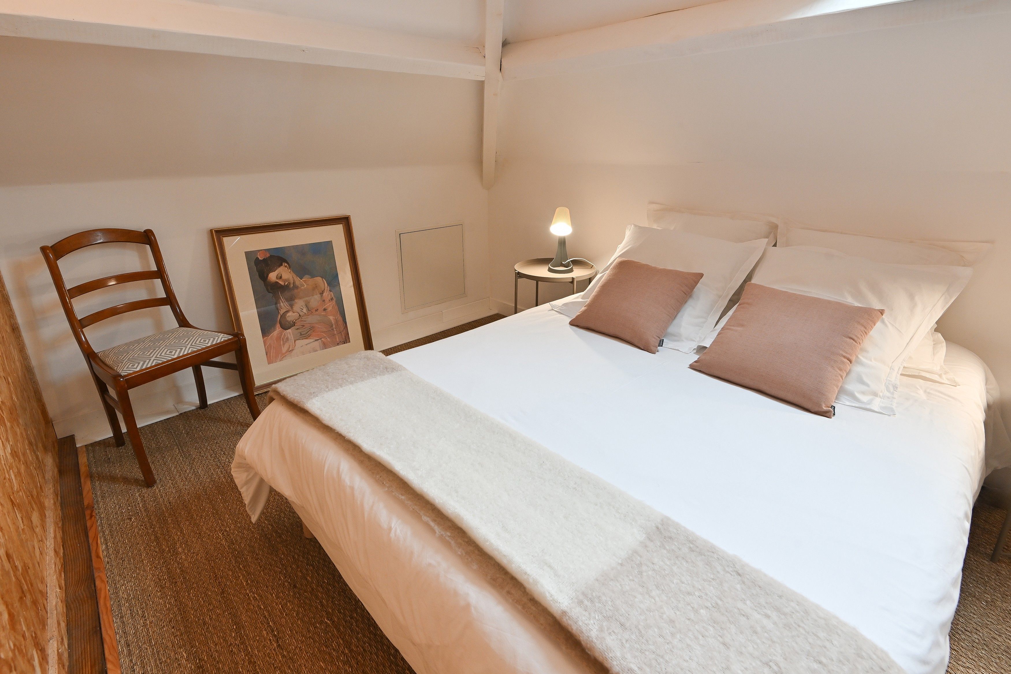 A neatly made bed with pillows and a blanket, a wooden chair, a small table with a lamp, and a framed picture on the wall.