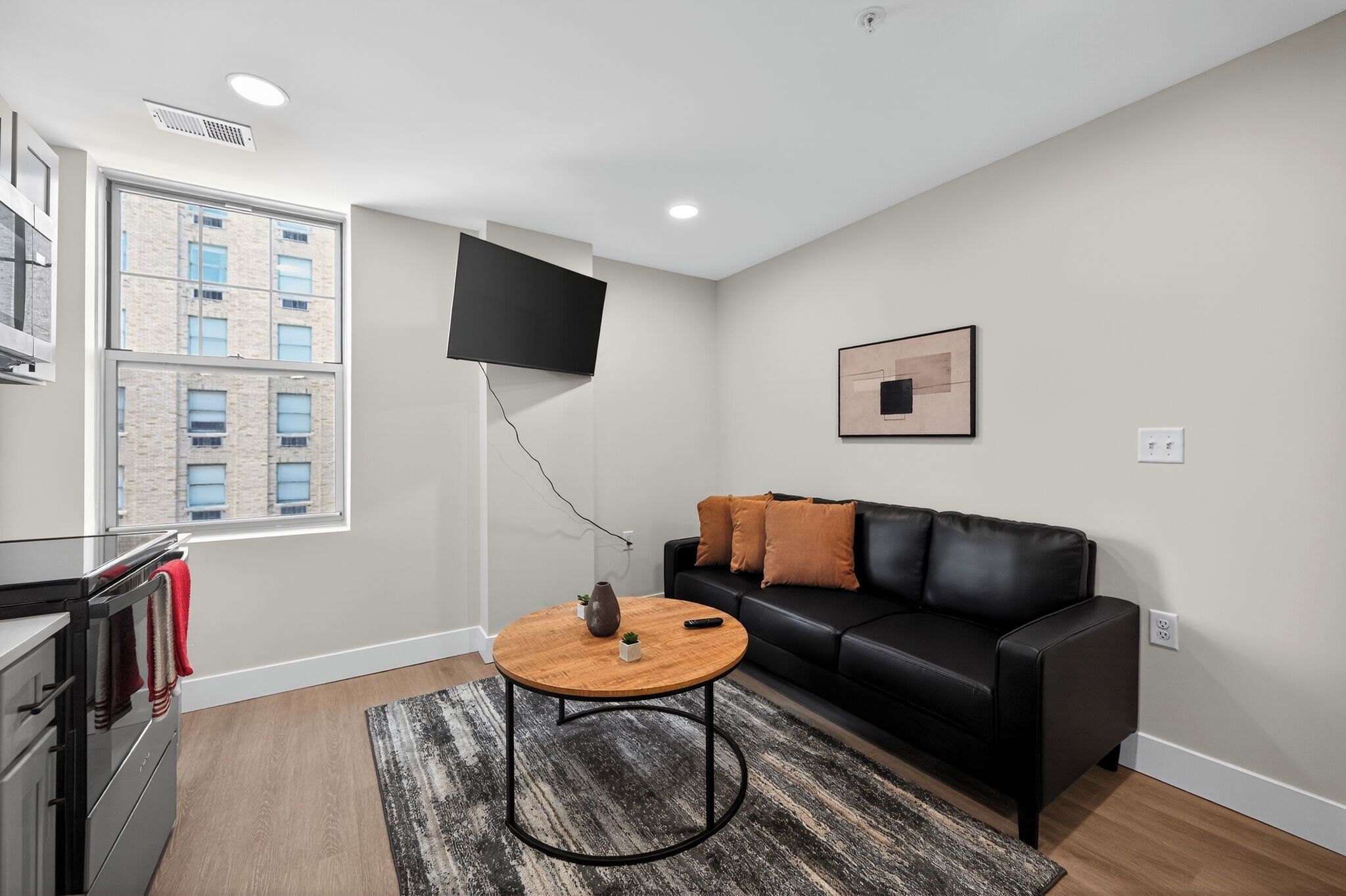 Un salon moderne avec un canapé en cuir noir, une table basse ronde en bois, un téléviseur à écran plat fixé au mur et une grande fenêtre donnant sur un immeuble.