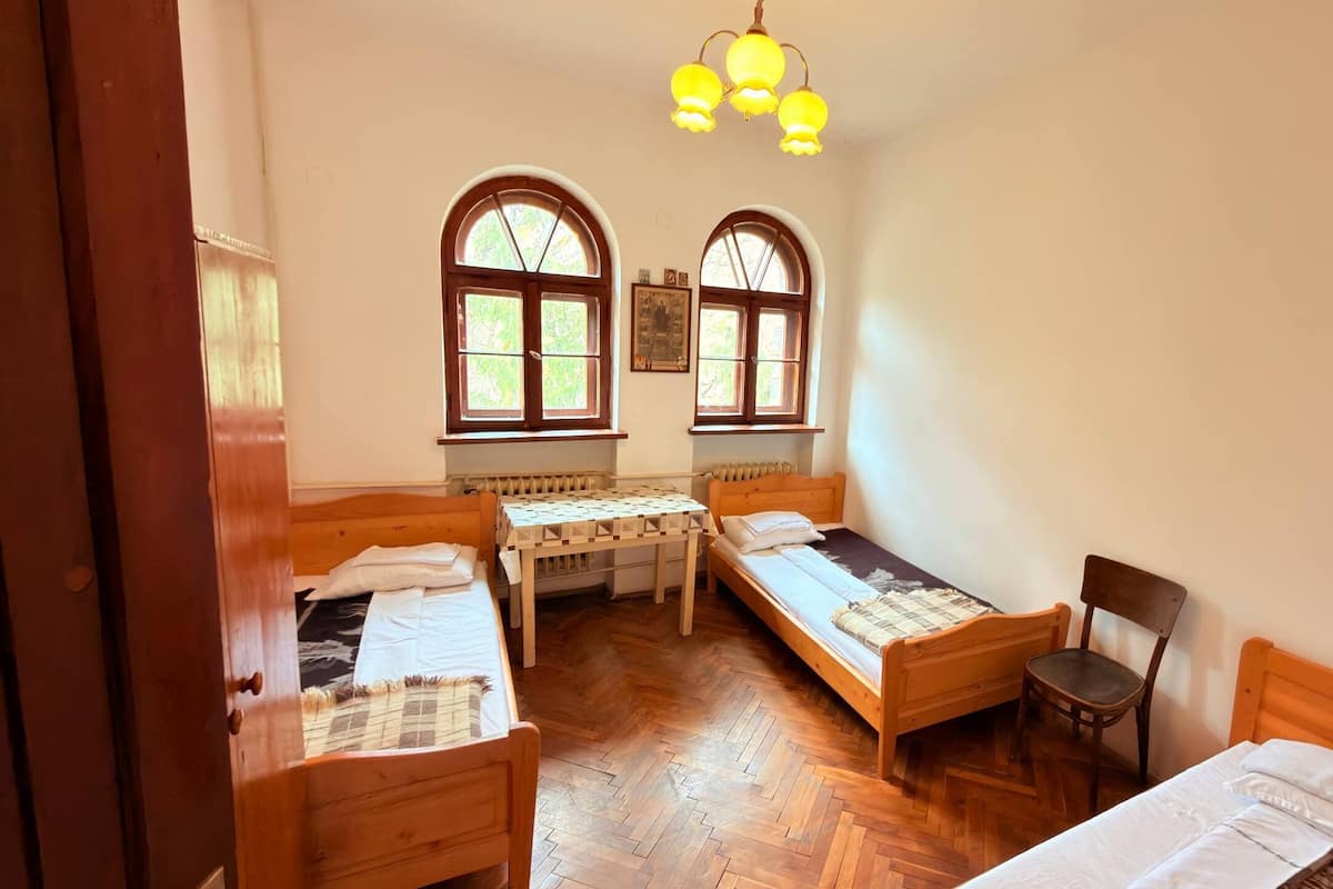 A room with two wooden beds, a small table, a chair, and two windows with curtains.