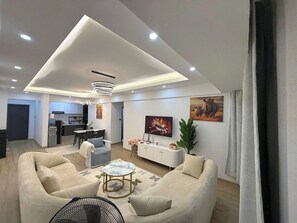 Ein modernes Wohnzimmer mit einer Couch, einem Couchtisch und einem Fernseher. Es gibt einen Essbereich mit Stühlen und einem Tisch, und im Hintergrund eine Küche.