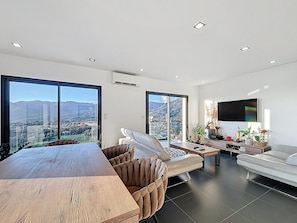 Una sala de estar moderna con una gran mesa de madera, un sofá, un televisor y ventanas amplias con vistas a la ciudad y a las montañas.
