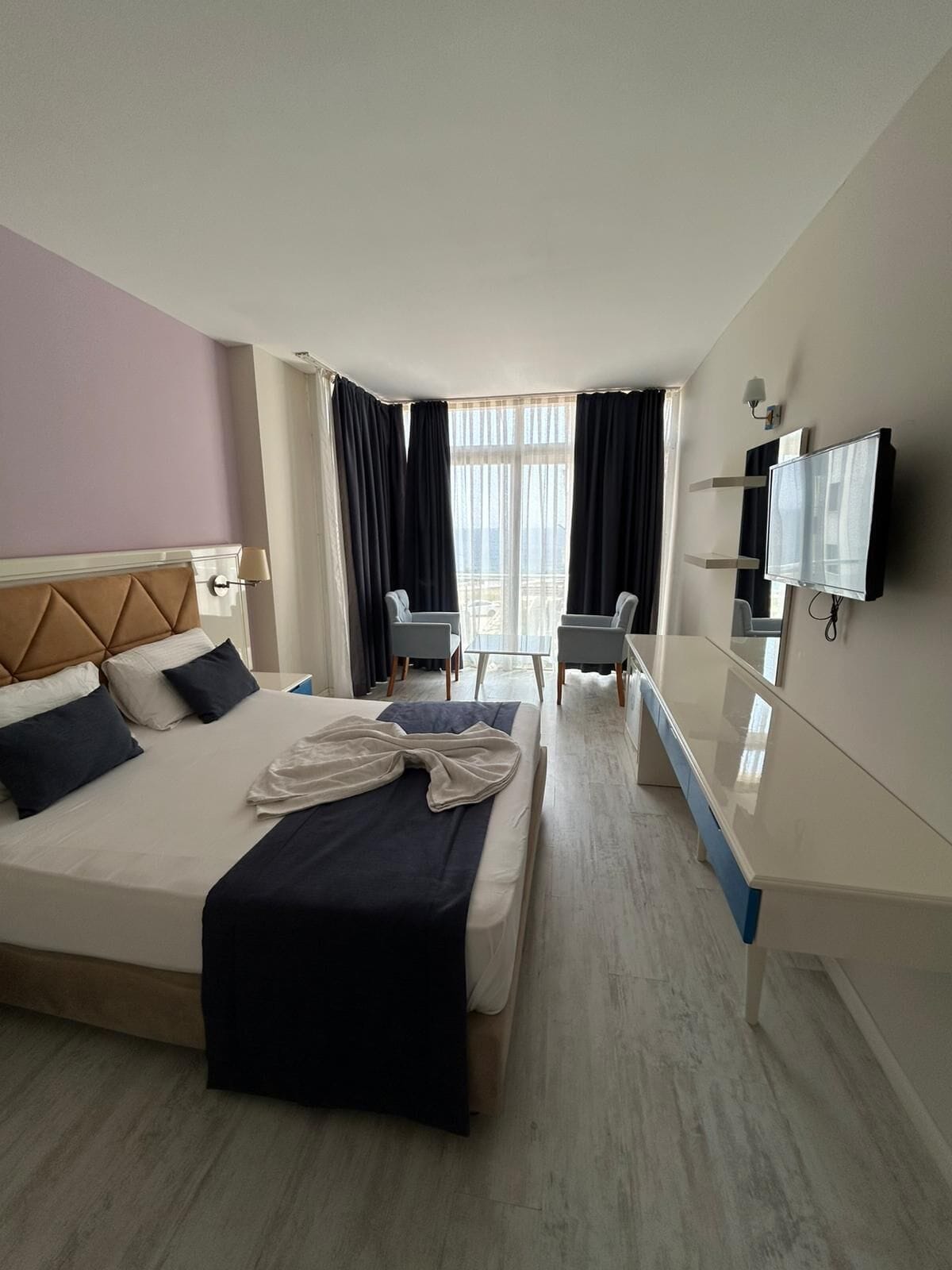 A hotel room with a bed, two armchairs, a TV, and a balcony view.