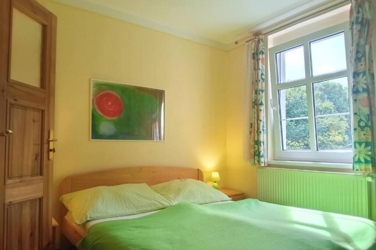 A room with a bed, a window with curtains, a door, and a wall painting.
