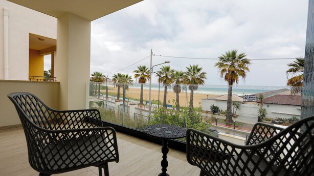 A balcony with two chairs and a table, overlooking a beach with palm trees and a clear sky. - Seõra Luxury Rooms (Porto Empedocle)