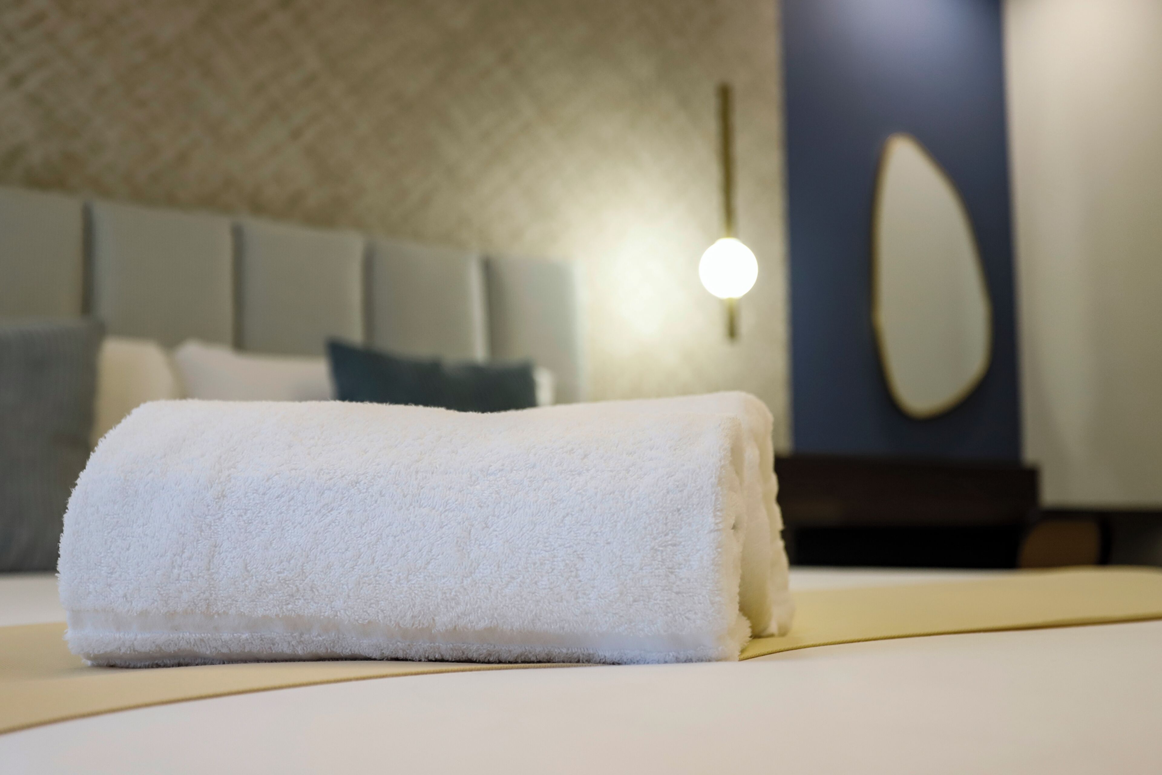 A neatly made bed with a rolled white towel, a headboard, and a wall-mounted light fixture.