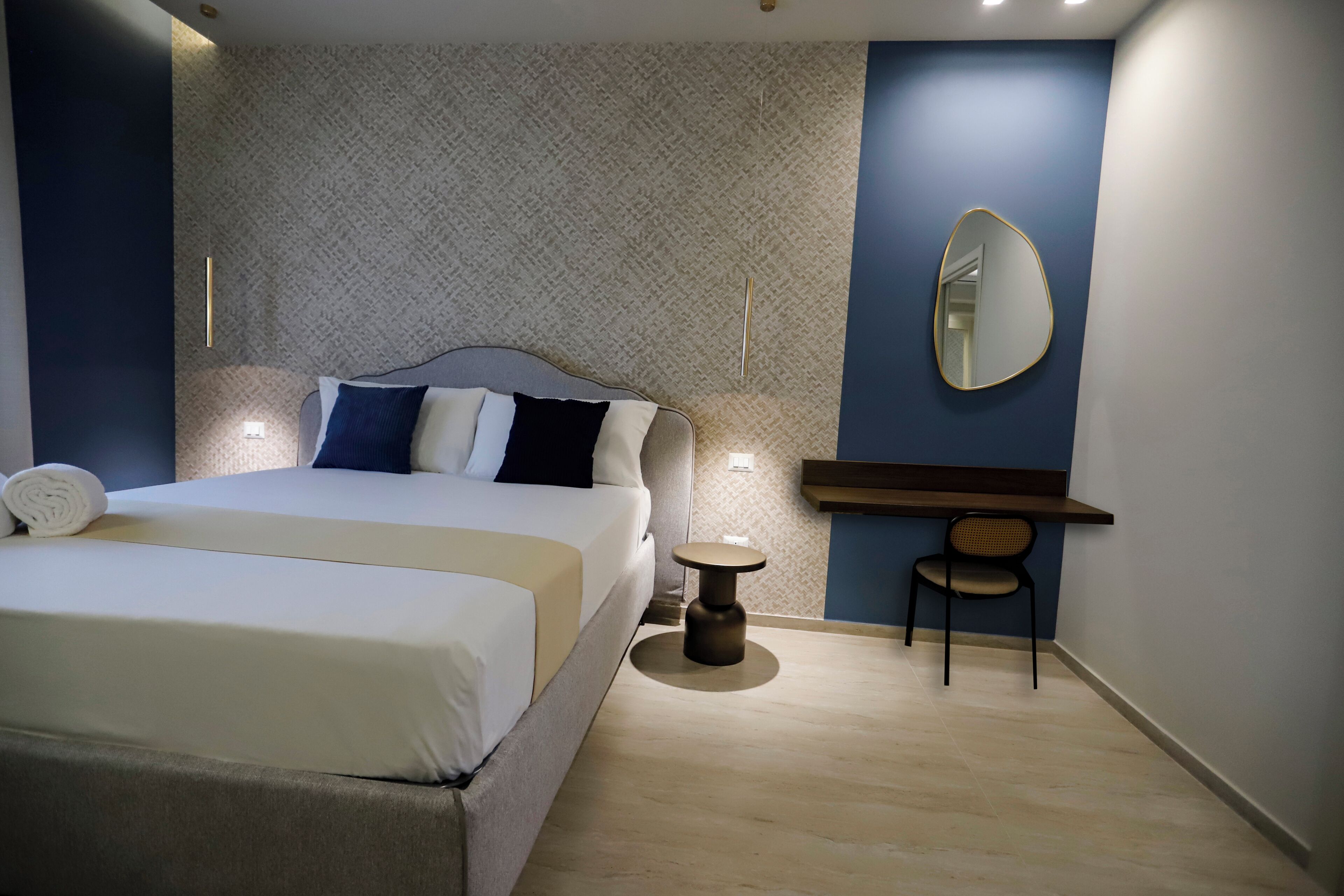 A modern hotel room with a neatly made bed, a small round table, a chair, a mirror, and a patterned wall.