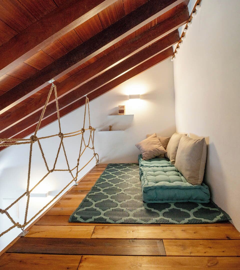 Een knus zolderkamer met een houten plafond, een hangende touwschommel, een groen patroonkleed en een blauwgroene bank met kussens.