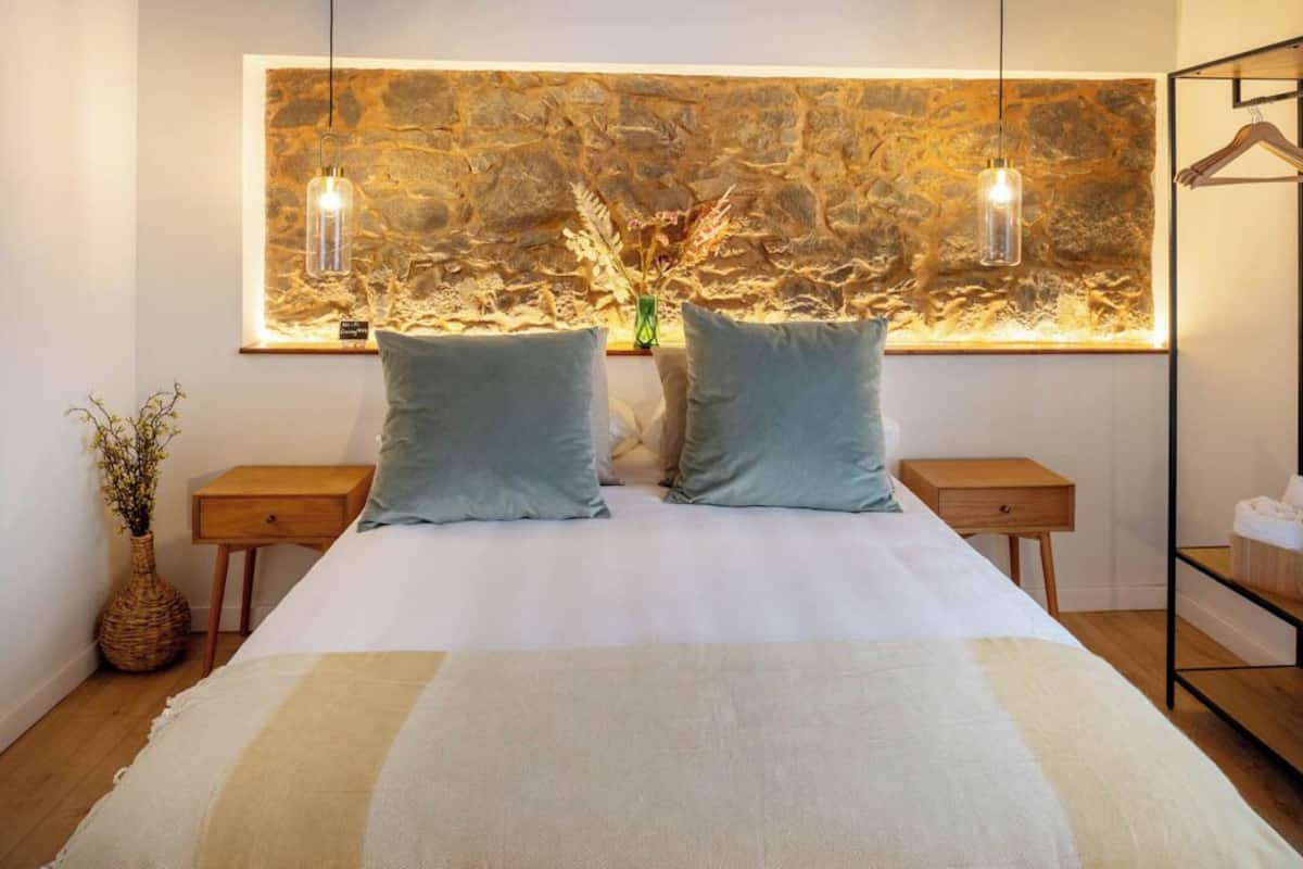 Una cama con dos almohadas, una pared de piedra, dos mesitas de noche, una lámpara colgante y un estante de madera.