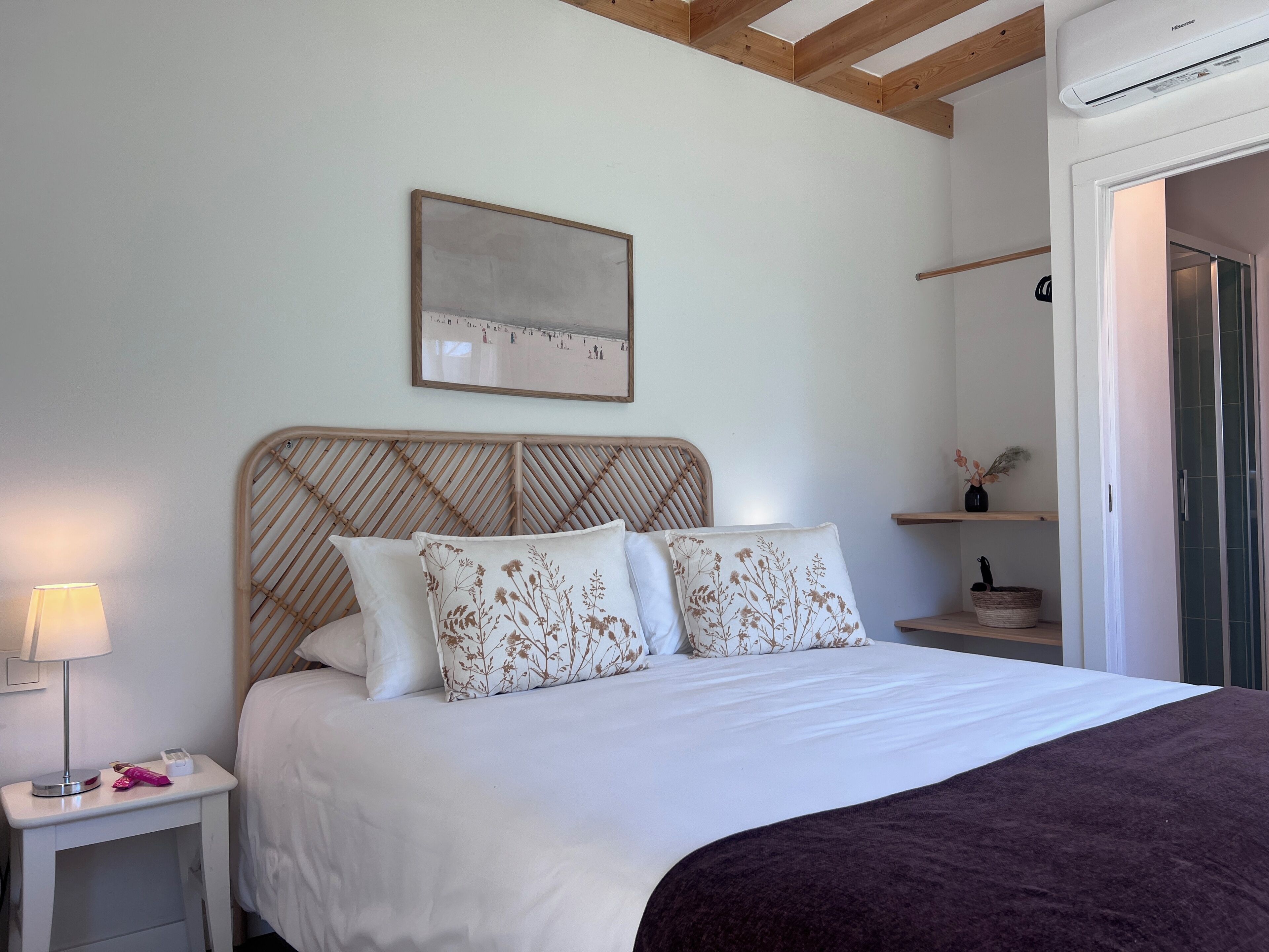 A bed with a wooden headboard, white bedding, and decorative pillows. There is a bedside table with a lamp and a small item on it. A framed picture hangs on the wall above the bed.