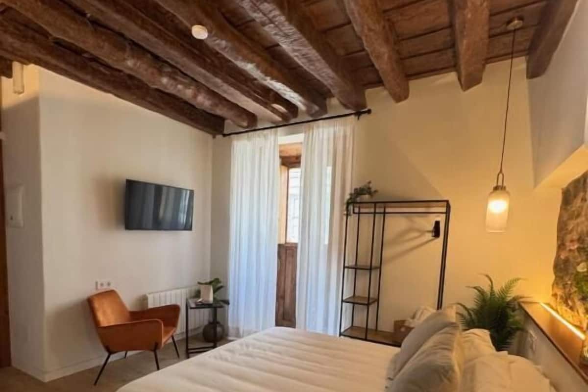 Un dormitorio con techo de madera, una cama con ropa blanca, una silla, un televisor y una planta.