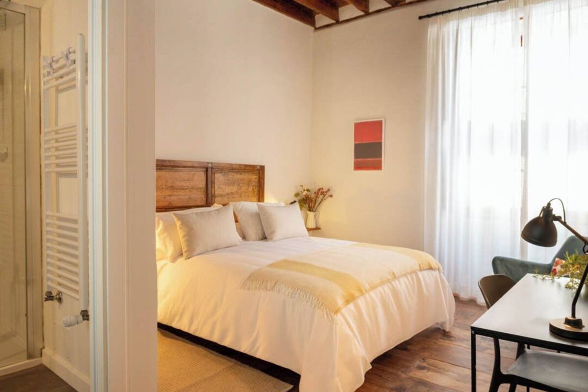 Un dormitorio con techo de madera, una cama con ropa de cama blanca, una mesita de noche con una lámpara y una ventana con cortinas.