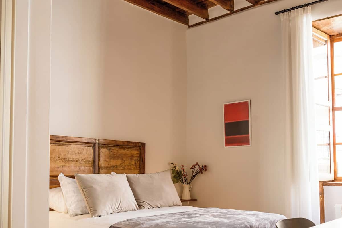 Un dormitorio con techo de madera, una cama con manta gris, pared blanca con un cuadro rojo y negro, y una ventana con cortinas.