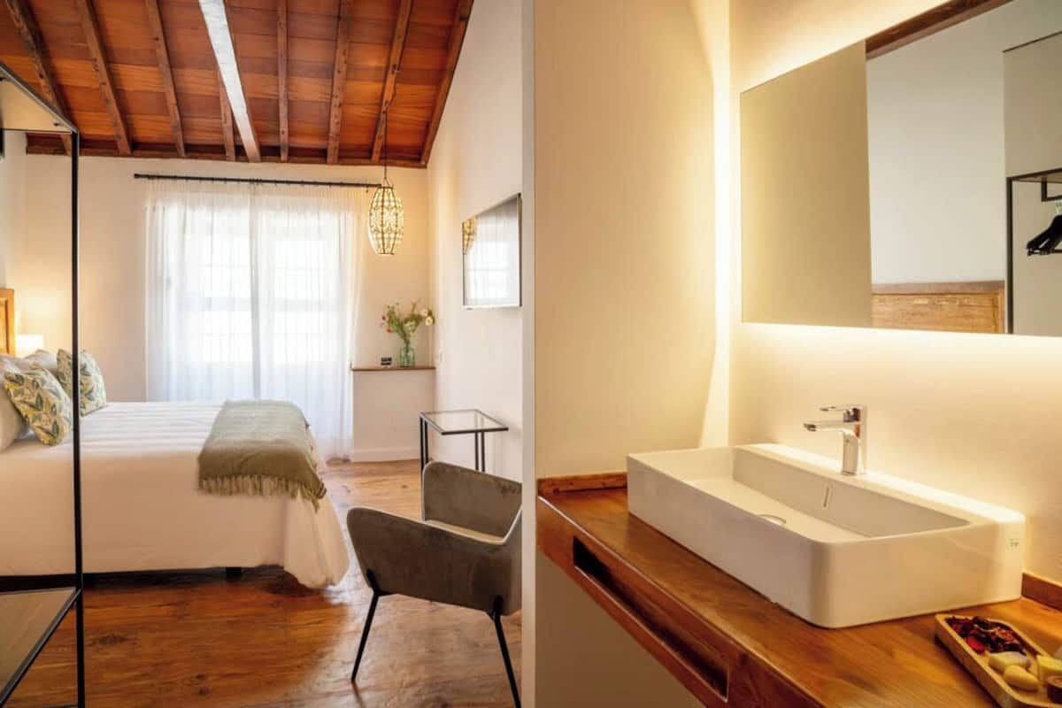 Un dormitorio con cama, una silla, una mesita de noche, una ventana con cortinas y techo de madera.