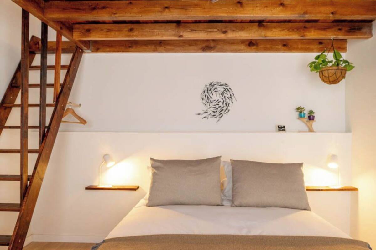 Un dormitorio con techo de madera, una cama con dos almohadas, una lámpara fijada a la pared, una planta pequeña y una escalera de madera.